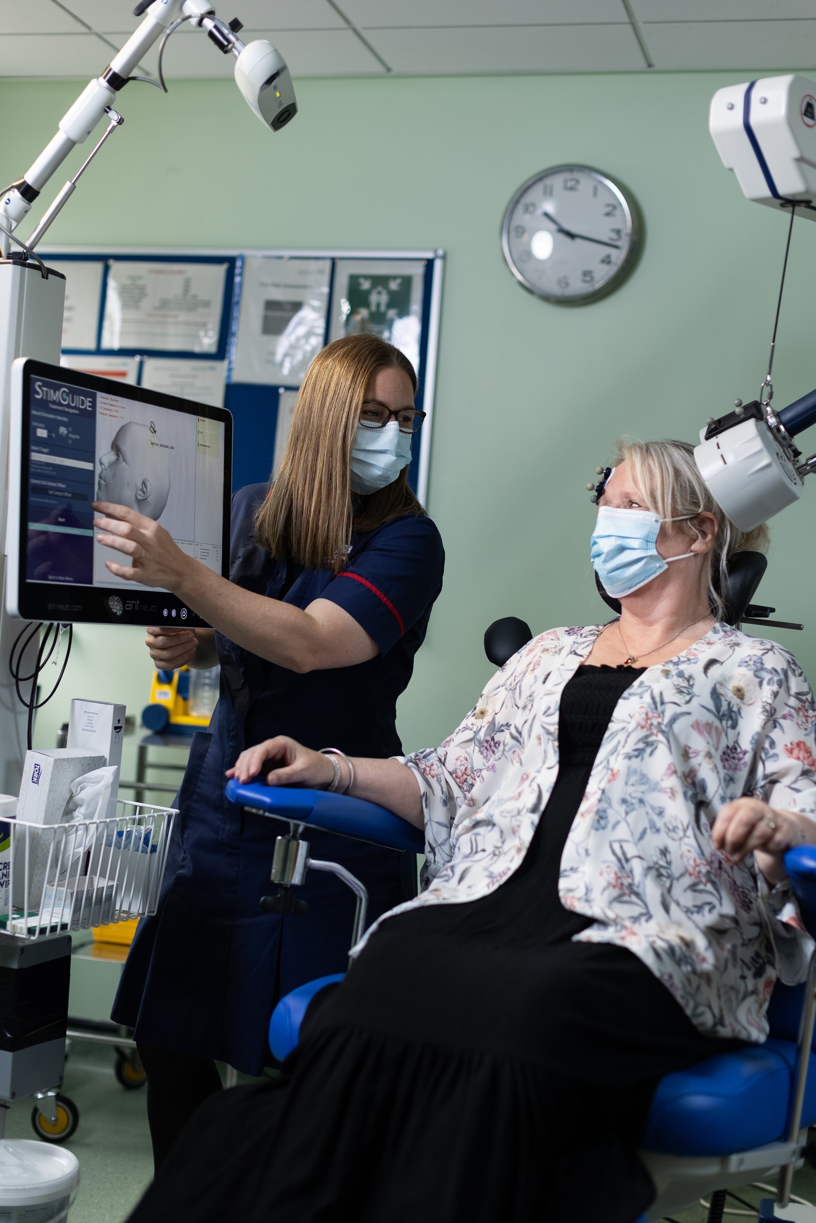 Woman sat in reclining chair with magnetic coils on her scalp, while nurse in uniform explains what's involved