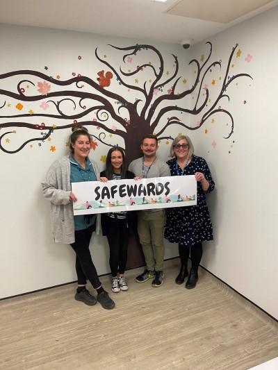 Four colleagues standing in front of a tree mural on Hope unit, holding a banner which says safewards